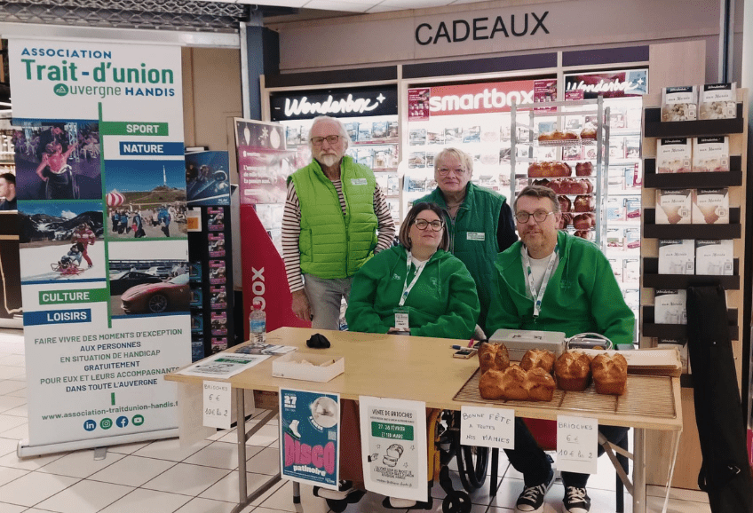 Dans un magasin Intermarché. 4 personnes souriantes de l'association dont une personne en fauteuil roulant manuel sont derrière une table. Sur la table des brioches sont proposées à la vente pour l'association.
