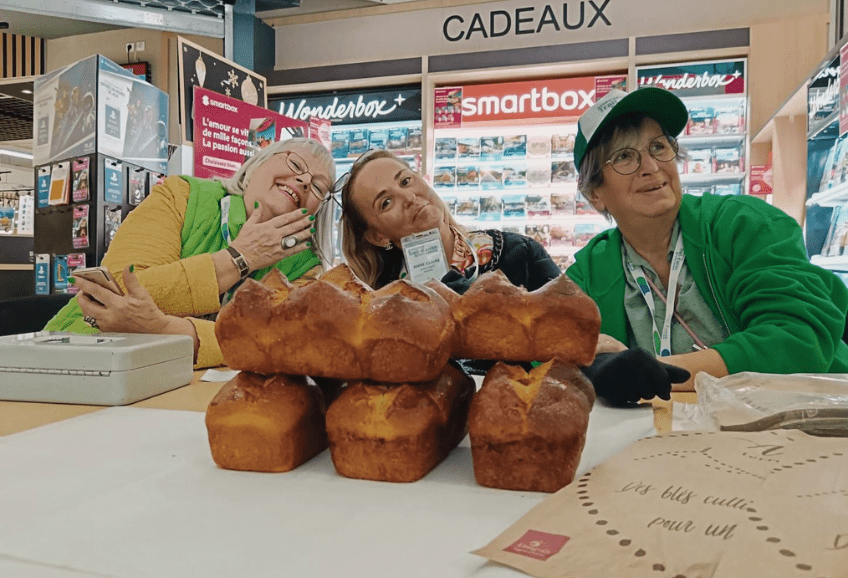 Dans un magasin Intermarché. 3 femmes souriantes de l'association sont derrière une table. Sur la table des brioches sont proposées à la vente pour l'association.