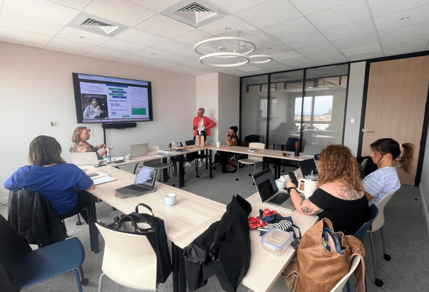 Jour 1 de Formation réalisée par Sandrine de l'association AcceSens au PIC Dans une salle de réunion où les tables forme un carré, Sandrine, Sandra et Marie de l'association Trait d'union Auvergne Handis sont assisent autour des tables et écoutent Orane et Aurélie également de trait d'union, qui présentent leur travail projeté sur un écran. Sandrine d'Accesens leur donne un retour sur leur travail.