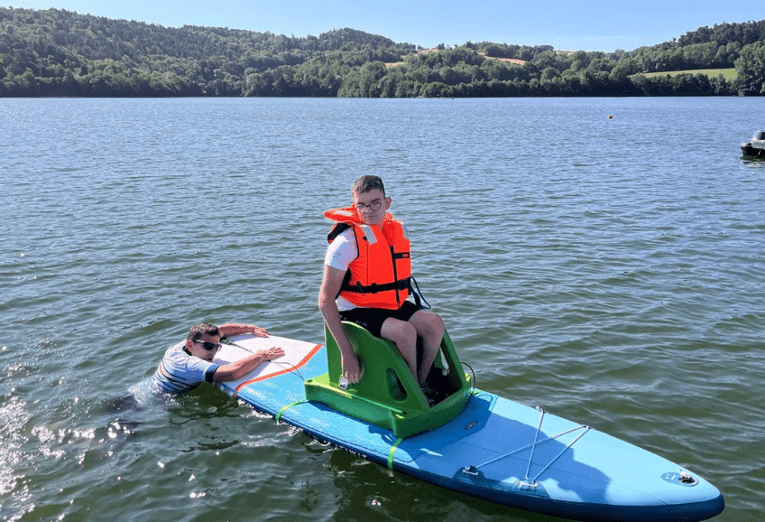 Un garçon en gilant orange de sauvetage assis sur un fauteuil fixé sur un paddle. Un homme dans l'eau assure la direction du paddle.