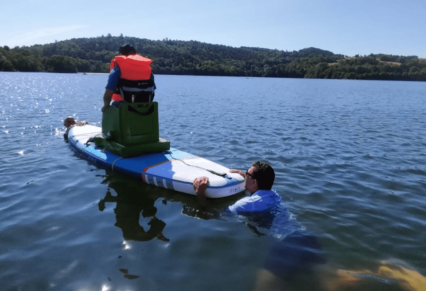 Un garçon en gilant orange de sauvetage assis sur un fauteuil fixé sur un paddle. deux hommes dans l'eau assurent la direction du paddle.