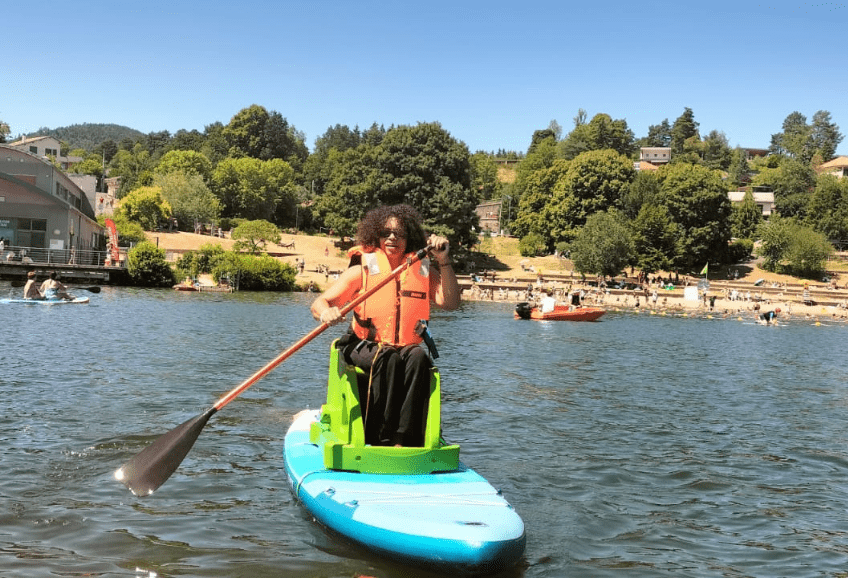 Une femme en situation de handicap assise sur un paddle adapté pagaie. Elle porte un gilet de sauvetage orange