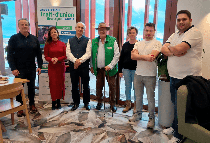 Remise Cheque Salaisons Bernard au profit de l'association. 7 personnes souriantes sont debout dans une salle du PIC locaux de l'association. Elles regardent en direction du photographe. Derrière Marie on peut voir le logo de l'Association Trait d'union Auvergne Handis